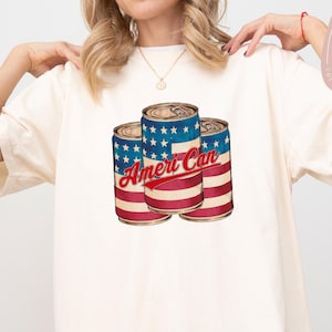 May include: Cream-colored t-shirt featuring a graphic of three beverage cans with a patriotic design. The cans have a blue starry top, red and white stripes, and the text "Ameri Can" in red. The shirt is a relaxed fit.