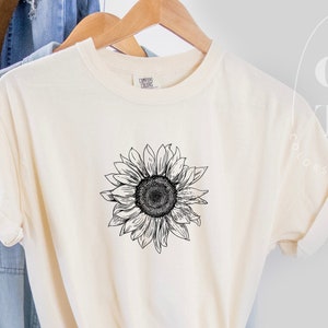 Puede incluir: Una camiseta blanca con un gr&aacute;fico de girasol en blanco y negro. La camiseta est&aacute; colgada de una percha y est&aacute; doblada por la mitad.