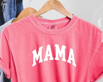 May include: A pink t-shirt with the word "MAMA" printed in white on the front. The shirt is hanging on a wooden hanger and is knotted at the bottom.