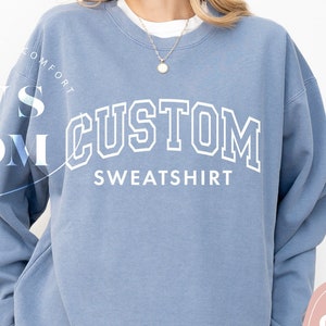 May include: A light blue sweatshirt with the text "CUSTOM SWEATSHIRT" in white outlined letters. The text is in an arch shape.