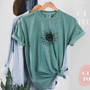 Puede incluir: Una camiseta azul turquesa con un gr&aacute;fico de girasol en blanco y negro. La camiseta est&aacute; hecha de un material suave y c&oacute;modo y tiene un corte holgado.