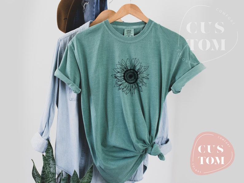 Puede incluir: Una camiseta azul turquesa con un gr&aacute;fico de girasol en blanco y negro. La camiseta est&aacute; hecha de un material suave y c&oacute;modo y tiene un corte holgado.
