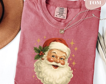 May include: A red-pink t-shirt featuring a vintage illustration of Santa Claus with a white beard and red hat. The design includes holly and gold stars. The shirt has a crew neck and a small tag that says "COMFORT COLORS". A silver necklace with a pendant is also visible.