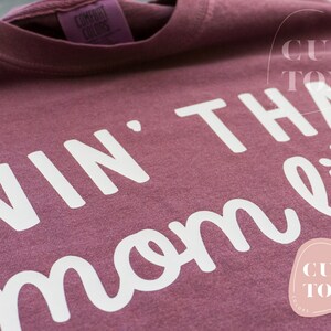 K&ouml;nnte beinhalten: Ein weinrotes Comfort Colors T-Shirt mit dem wei&szlig;en Schriftzug "Livin' That Mom Life". Das Shirt hat ein Etikett mit der Aufschrift "Comfort Colors". Ein individuelles Logo ist sichtbar.