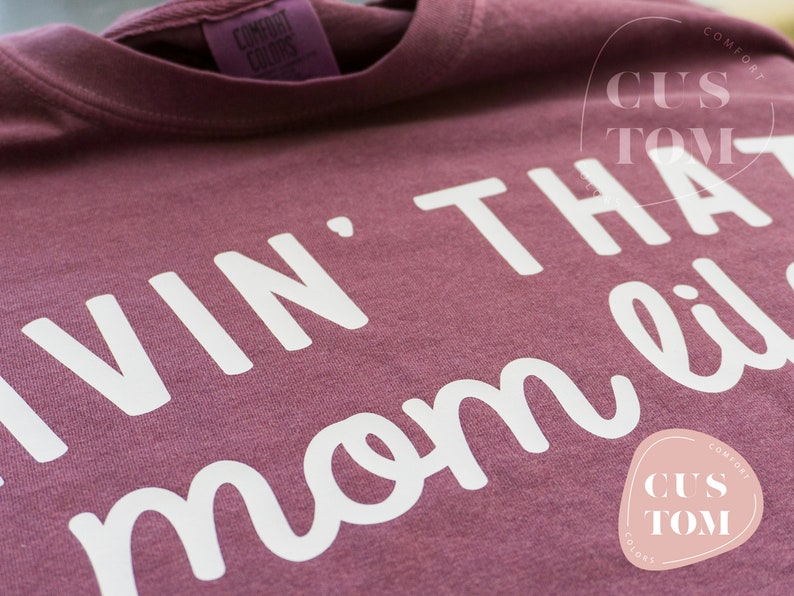 K&ouml;nnte beinhalten: Ein weinrotes Comfort Colors T-Shirt mit dem wei&szlig;en Schriftzug "Livin' That Mom Life". Das Shirt hat ein Etikett mit der Aufschrift "Comfort Colors". Ein individuelles Logo ist sichtbar.
