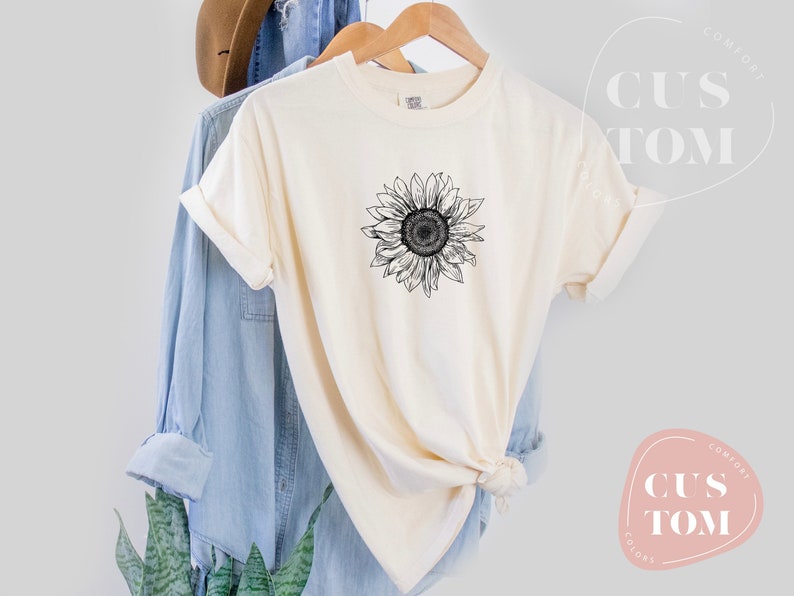 Puede incluir: Una camiseta blanca con un gr&aacute;fico de girasol en blanco y negro. La camiseta est&aacute; colgada de una percha y est&aacute; doblada por la mitad.