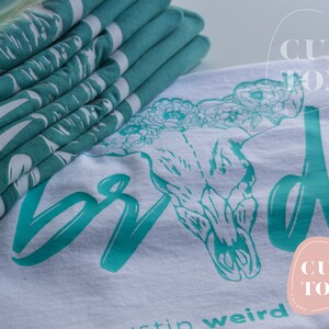 Op de afbeelding: Een wit T-shirt met een turquoise schedel- en bloemenontwerp. De tekst "Bride" is in turquoise over de voorkant van het shirt geschreven. Het shirt heeft ook de tekst "Custom Comfort" in een turquoise cirkel.