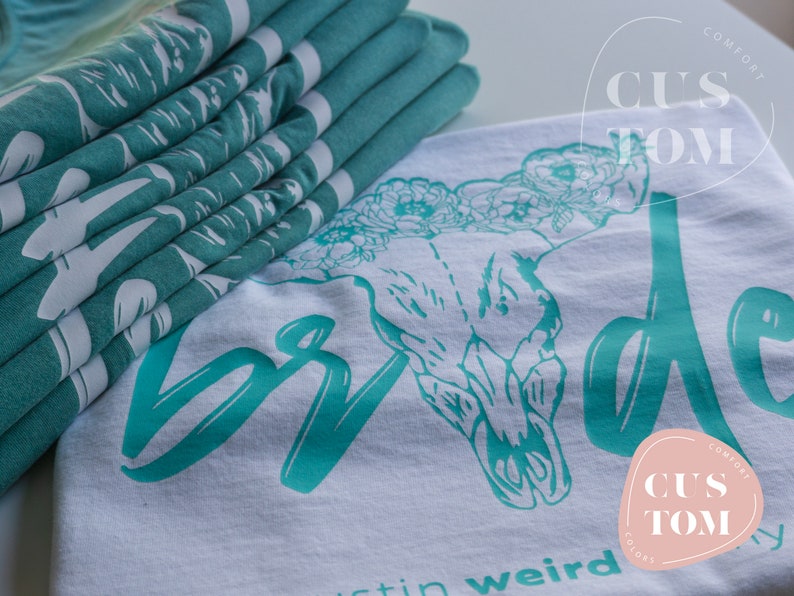 Op de afbeelding: Een wit T-shirt met een turquoise schedel- en bloemenontwerp. De tekst "Bride" is in turquoise over de voorkant van het shirt geschreven. Het shirt heeft ook de tekst "Custom Comfort" in een turquoise cirkel.