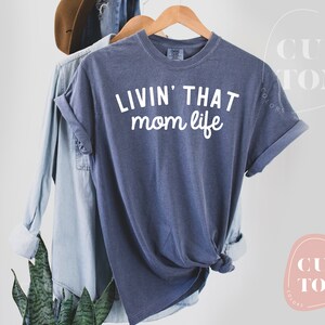 K&ouml;nnte beinhalten: Marineblaues T-Shirt mit dem wei&szlig;en Schriftzug "LIVIN' THAT mom life". Das T-Shirt h&auml;ngt an einem Holzb&uuml;gel, im Hintergrund befinden sich ein Jeanshemd und ein brauner Hut. Die &Auml;rmel des T-Shirts sind hochgekrempelt.