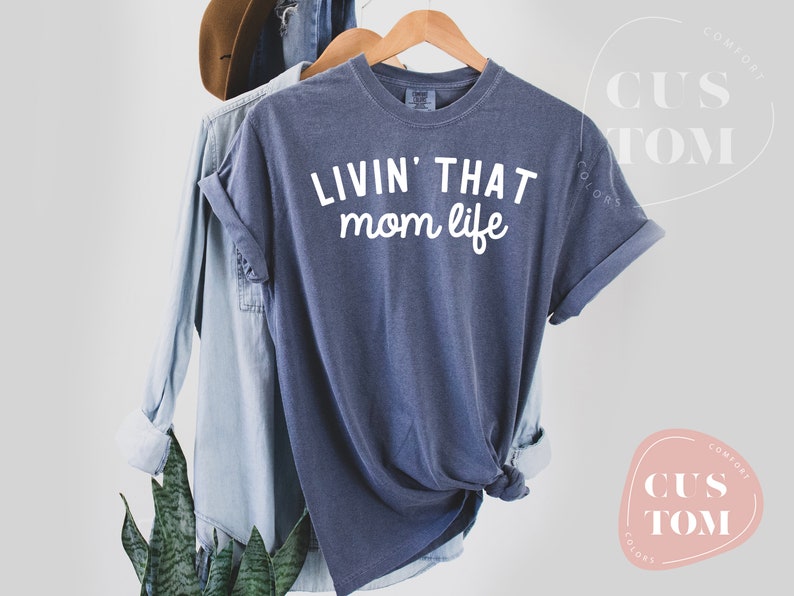 K&ouml;nnte beinhalten: Marineblaues T-Shirt mit dem wei&szlig;en Schriftzug "LIVIN' THAT mom life". Das T-Shirt h&auml;ngt an einem Holzb&uuml;gel, im Hintergrund befinden sich ein Jeanshemd und ein brauner Hut. Die &Auml;rmel des T-Shirts sind hochgekrempelt.