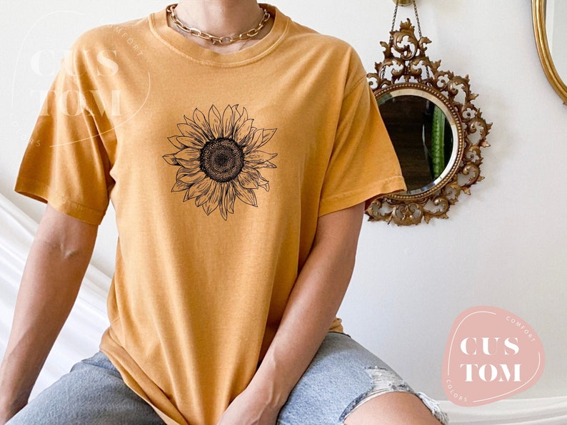Puede incluir: Una camiseta de color amarillo mostaza con un gr&aacute;fico de girasol en blanco y negro. La camiseta tiene cuello redondo y mangas cortas.