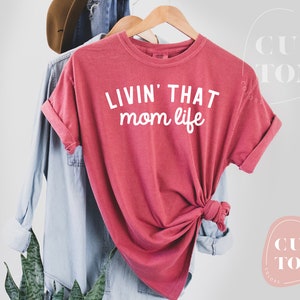 K&ouml;nnte beinhalten: Ein rot-rosa T-Shirt mit dem wei&szlig;en Schriftzug "LIVIN' THAT mom life". Das Shirt h&auml;ngt an einem Holzb&uuml;gel mit einem hellblauen Hemd und einem braunen Hut im Hintergrund.