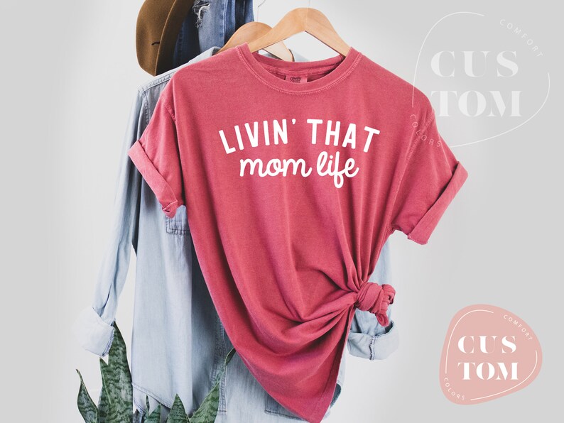 K&ouml;nnte beinhalten: Ein rot-rosa T-Shirt mit dem wei&szlig;en Schriftzug "LIVIN' THAT mom life". Das Shirt h&auml;ngt an einem Holzb&uuml;gel mit einem hellblauen Hemd und einem braunen Hut im Hintergrund.