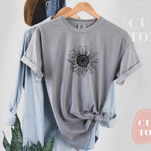 Puede incluir: Una camiseta gris con un dibujo de l&iacute;nea negra de un girasol en la parte delantera. La camiseta est&aacute; doblada y colgada en una percha.