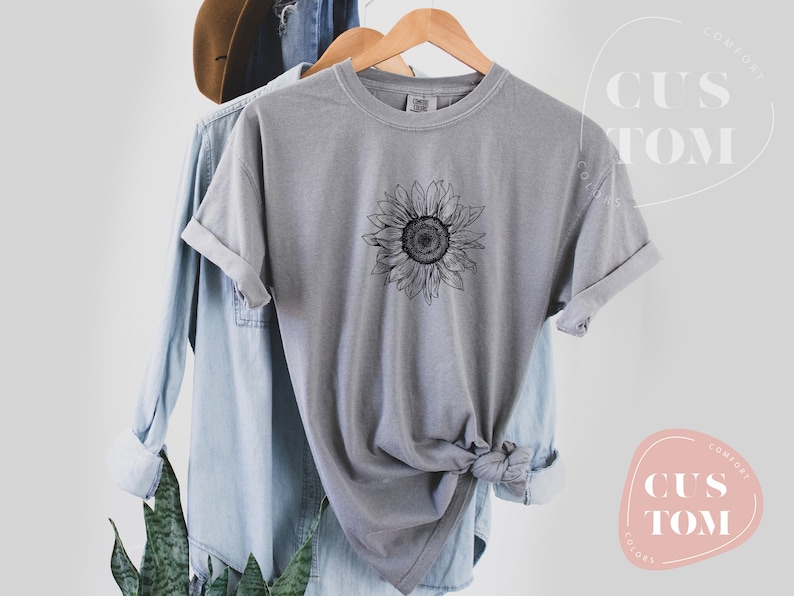 Puede incluir: Una camiseta gris con un dibujo de l&iacute;nea negra de un girasol en la parte delantera. La camiseta est&aacute; doblada y colgada en una percha.