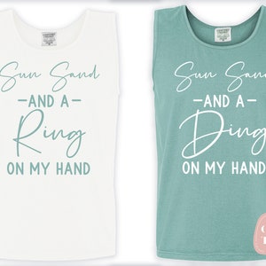 Op de afbeelding: Twee witte tanktops met een teal blauwe grafische afbeelding met de tekst "Sun Sand -AND A- Ring ON MY HAND" en "Sun Sand -AND A- Ding ON MY HAND".