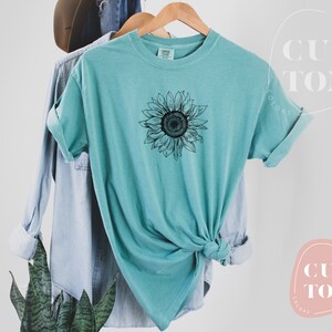 Puede incluir: Una camiseta azul turquesa con un gr&aacute;fico de girasol en blanco y negro. La camiseta est&aacute; anudada en la parte inferior.