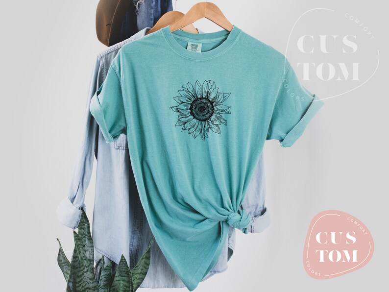 Puede incluir: Una camiseta azul turquesa con un gr&aacute;fico de girasol en blanco y negro. La camiseta est&aacute; anudada en la parte inferior.