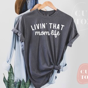 K&ouml;nnte beinhalten: Dunkelgraues T-Shirt mit dem wei&szlig;en Aufdruck "LIVIN' THAT mom life". Das Shirt h&auml;ngt an einem Holzb&uuml;gel, im Hintergrund sind ein hellblaues Hemd und ein brauner Hut zu sehen. Das T-Shirt ist am unteren Saum geknotet.