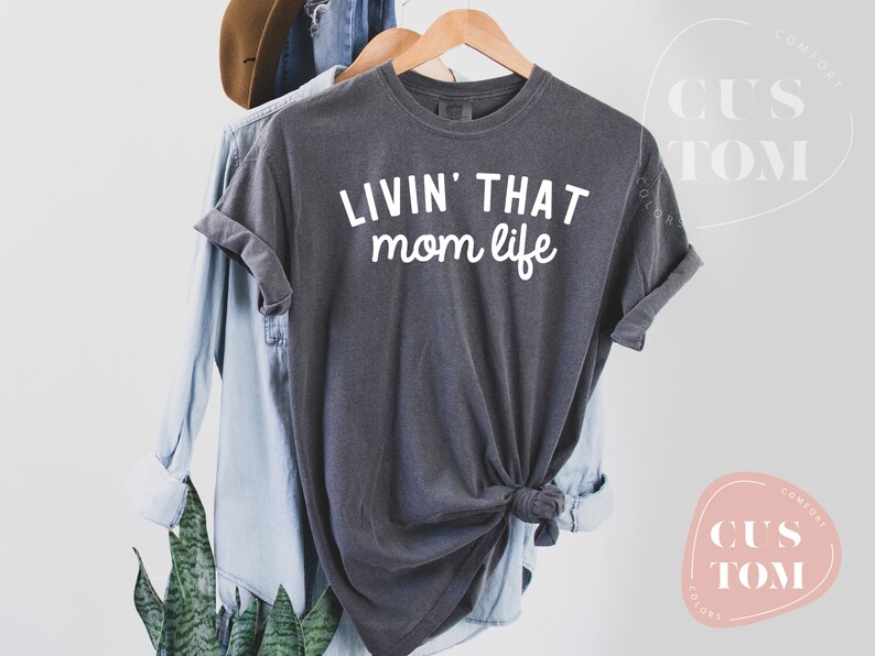 K&ouml;nnte beinhalten: Dunkelgraues T-Shirt mit dem wei&szlig;en Aufdruck "LIVIN' THAT mom life". Das Shirt h&auml;ngt an einem Holzb&uuml;gel, im Hintergrund sind ein hellblaues Hemd und ein brauner Hut zu sehen. Das T-Shirt ist am unteren Saum geknotet.