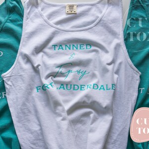 Op de afbeelding: Drie witte tanktops met de tekst "Tanned & Tipsy Fort Lauderdale" in turquoise gedrukt op de voorkant. De tanktops zijn in een driehoekige vorm gerangschikt, met het bovenste tanktop in het midden en de andere twee tanktops aan weerszijden.