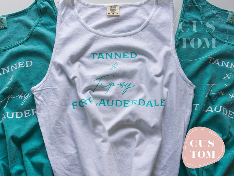 Op de afbeelding: Drie witte tanktops met de tekst "Tanned & Tipsy Fort Lauderdale" in turquoise gedrukt op de voorkant. De tanktops zijn in een driehoekige vorm gerangschikt, met het bovenste tanktop in het midden en de andere twee tanktops aan weerszijden.