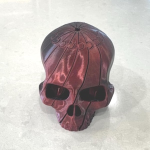 Op de afbeelding: Een rood en zwart schedelvormig decoratief object. De schedel heeft grote oogkassen, een prominente neusholte en een gedetailleerde bovenkant met verticale lijnen. De kleur gaat over van rood naar zwart aan de bovenkant.