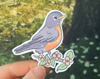 May include: A sticker featuring a cartoon robin perched on a flowering branch. The bird has gray feathers, an orange breast, and a yellow beak. The branch has pink flowers and green leaves. The sticker is held against a blurred green background.