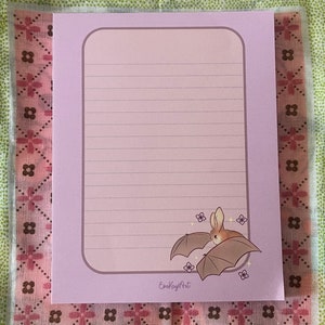 May include: A purple lined notepad with a pink border and a cute cartoon bat illustration. The notepad has the text "EmKayArt" at the bottom.