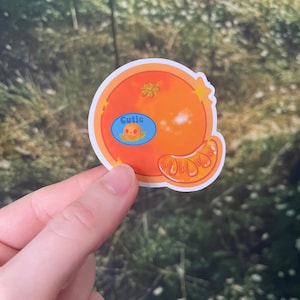 May include: A sticker of a cartoon orange with a blue circle that says "Cutie" and a small cartoon face inside the circle. The orange is a bright orange color and has a small piece of peel on the side.