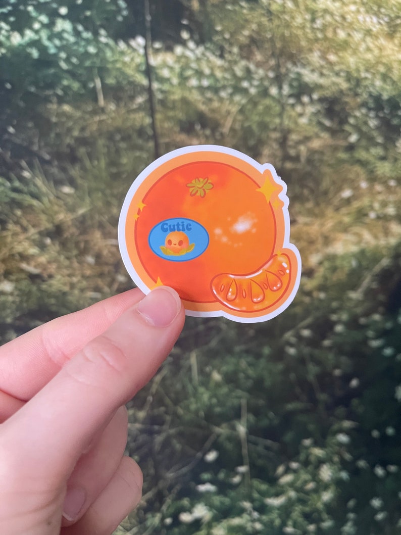 May include: A sticker of a cartoon orange with a blue circle that says "Cutie" and a small cartoon face inside the circle. The orange is a bright orange color and has a small piece of peel on the side.