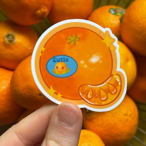 May include: A sticker of a cartoon orange with a smiling face and the word "Cutie" in a blue speech bubble. The orange is surrounded by real oranges in a basket.