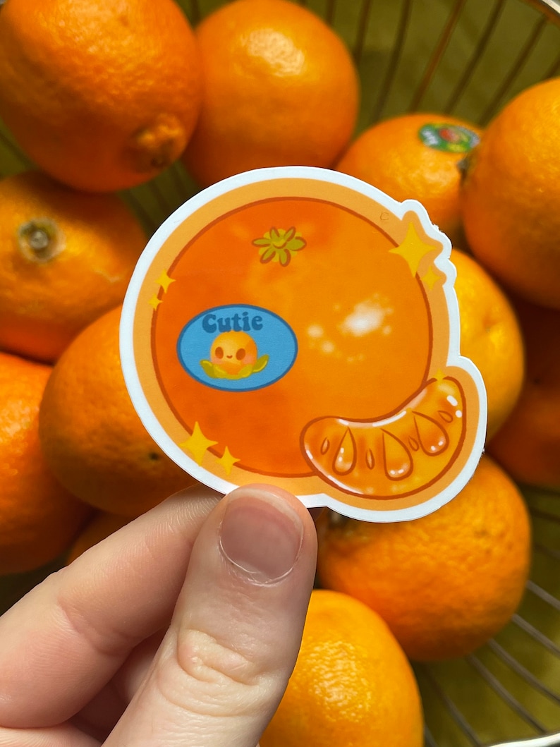 May include: A sticker of a cartoon orange with a smiling face and the word "Cutie" in a blue speech bubble. The orange is surrounded by real oranges in a basket.