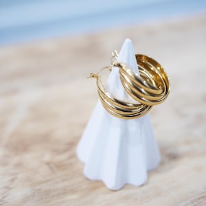 Peut inclure: Boucles d'oreilles créoles dorées à plusieurs niveaux, présentées sur un présentoir à bijoux blanc géométrique. Les boucles d'oreilles sont posées sur un fond bleu clair et imitation bois. Un bijou classique.