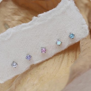 Może przedstawiać: Pięć małych srebrnych kolczyków z różnokolorowymi kamieniami szlachetnymi. Kamienie szlachetne są niebieskie, jasnoniebieskie, różowe, fioletowe i przezroczyste.