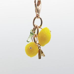 May include: A gold-toned metal charm featuring two bright yellow lemon-shaped pendants with a small green leaf accent and a single gold-toned letter A charm, all suspended from a matching gold-toned lobster claw clasp for easy attachment.
