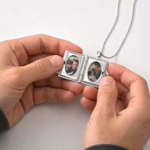 Puede incluir: Un collar con un relicario plateado con dos marcos de fotos ovalados. El relicario está abierto, revelando dos fotografías en color. La cadena del collar también es plateada. El relicario está sostenido por una persona.