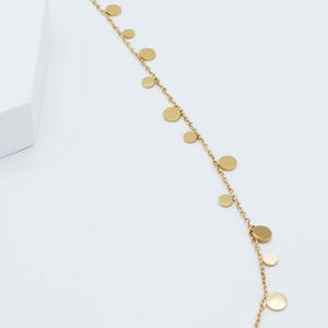 May include: A gold-toned chain necklace featuring a series of small, flat, circular discs spaced evenly along the length of the delicate metal links. The necklace is arranged in a gentle curve against a plain, light-colored background.