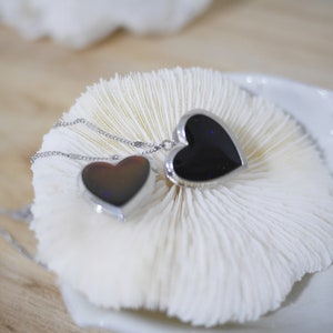 Puede incluir: Dos collares con colgantes en forma de corazón. Los colgantes tienen un centro negro con un borde plateado. Los collares están sobre una superficie blanca y texturizada. Las cadenas son plateadas y delicadas.