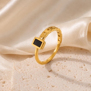 May include: A gold-toned ring featuring a rectangular black inlay set within a frame. The band of the ring displays a series of small, square-shaped cutouts along its upper half, creating a geometric pattern that leads to the central black stone.