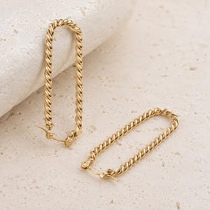 May include: A pair of gold-toned hoop earrings featuring an elongated oval shape with a twisted rope-like texture. The earrings have a hinged closure mechanism at the base, designed for secure attachment, and exhibit a polished metallic finish.