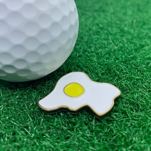 Puede incluir: Una bola de golf blanca con un marcador de bola de golf de huevo frito dorado y blanco sobre césped artificial verde.