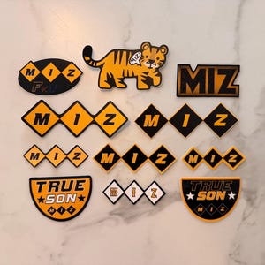 May include: A collection of patches and stickers with a black, gold, and white color scheme. Designs include the text "MIZ", a tiger, and the words "TRUE SON". The patches and stickers are in various shapes.