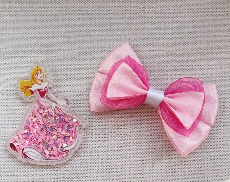 May include: A pink and white bow with a princess character on a clear background. The princess is wearing a pink dress with glitter.
