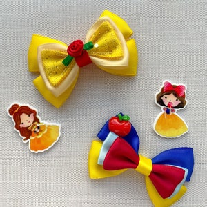 May include: Two hair bows featuring Disney princesses. One bow is yellow with a red rose and glitter. The other bow is red, blue, and yellow with a red apple charm.