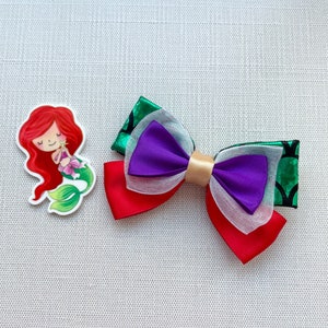 May include: A red, white, purple, and green bow with a gold center. The bow is decorated with a mermaid illustration.
