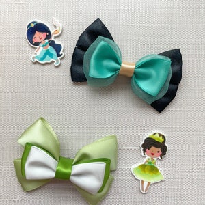 May include: Two green and white satin ribbon bows and two character hair clips. One clip features a cartoon of Jasmine from Aladdin, the other a cartoon of a princess with a frog.