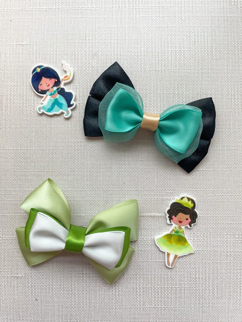 May include: Two green and white satin ribbon bows and two character hair clips. One clip features a cartoon of Jasmine from Aladdin, the other a cartoon of a princess with a frog.