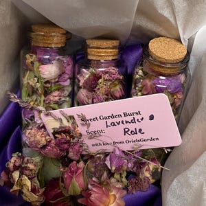 May include: Three small glass jars filled with dried lavender and other purple flowers. A pink card with the text "Sweet Garden Burst Scent: Lavender Role with love from OrielsGarden" is placed on top of the jars.