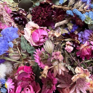 May include: A close-up of a colourful collection of dried flowers. The arrangement includes deep reds, purples, pinks, blues, and green accents. The composition is dense, highlighting the textures and forms of the dried petals and stems.